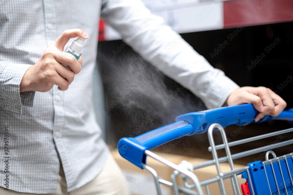 Male hand customer spraying hand sanitizer or alcohol gel on shopping cart while shopping in grocery store or supermarket. Cleaning and preventing COVID-19 (Coronavirus) infection. New normal concept