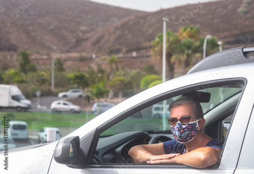 Senior woman wearing a flowered surgical mask due to the covid19 coronavirus parked with his silver car leaning against the window - active retired people concept