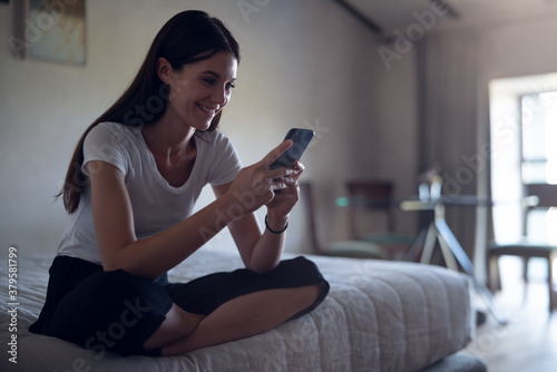 Authentic shot of an young woman is sitting on a bed and using a smart phone for send messages, make calls or navigate in internet just waked in bedroom at home.