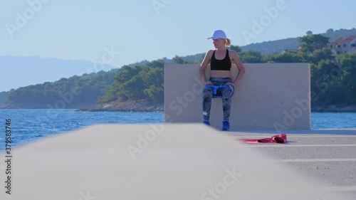A young blonde woman in a white cap exercises outside by the sea. Wall sit exercise. A beautiful summer morning.
