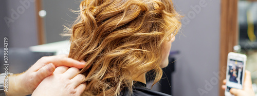 Hairdresser doing hairstyle for young woman with red curly hair and with smartphone in her hands in beauty salon