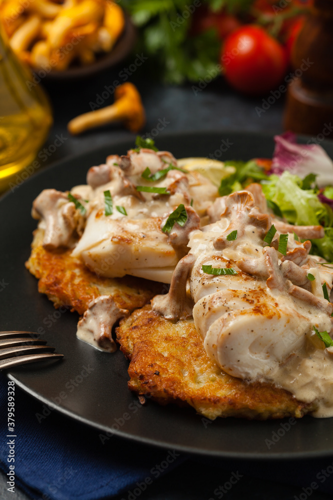 Fried cod, served on potato pancakes with mushrooms sauce and salads. Portion on a black plate. Dark background.