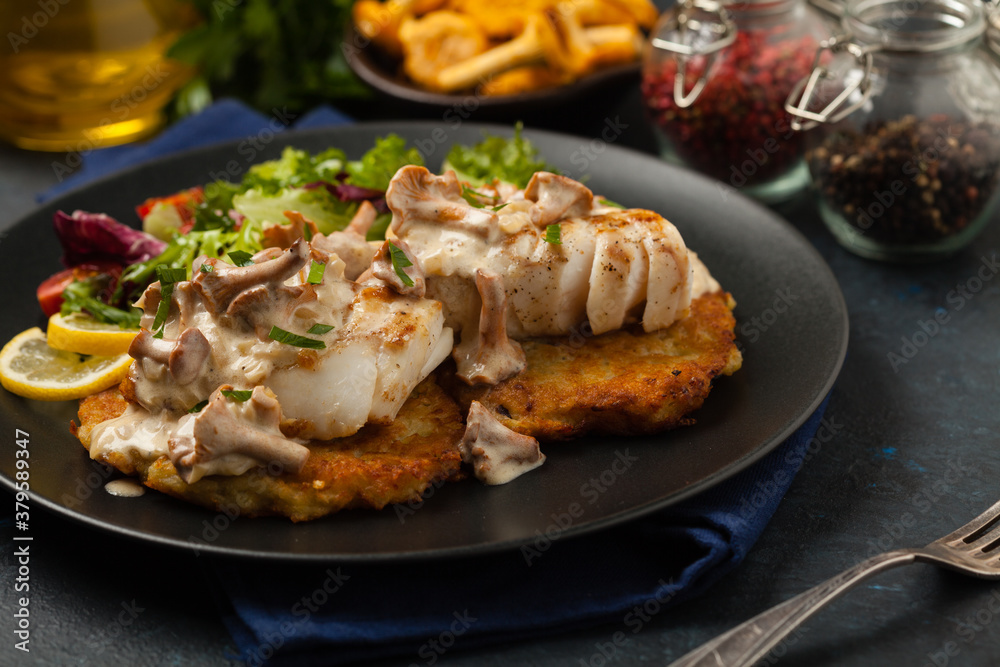 Fried cod, served on potato pancakes with mushrooms sauce and salads. Portion on a black plate. Dark background. Fried cod, served on potato pancakes with mushrooms sauce and salads. Portion on a black plate. Dark background.