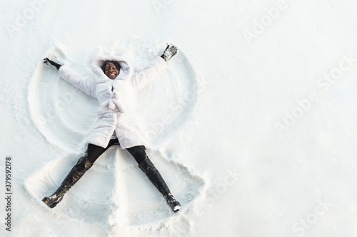 black girl in fur coat and high top boots lies on white glade and draws snow angel picture with arms and legs top view photo