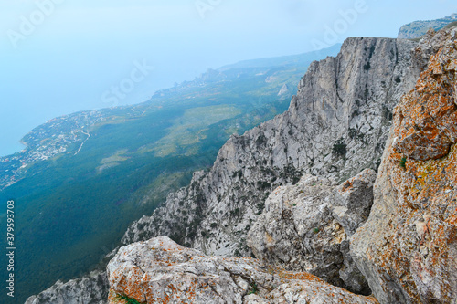 large steep sharp stone rock cliff with red moss against the background of Black sea coastline, the city and forest below, summer