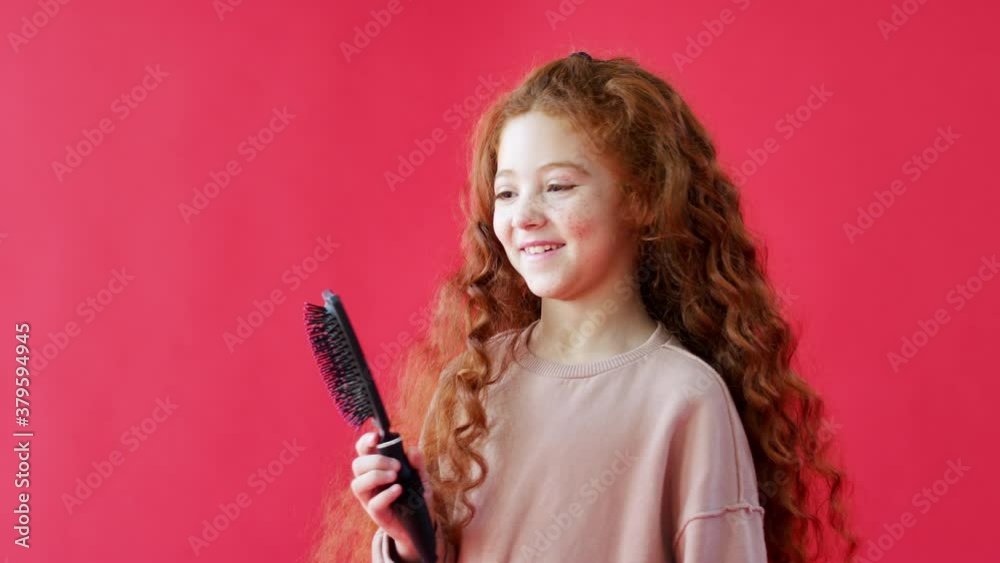 Young casually dressed girl with long red hair stands against red studio background holding hairbrush smiling and laughing - shot in slow motion Young casually dressed girl with long red hair stands against red studio background holding hairbrush smiling and laughing - shot in slow motion