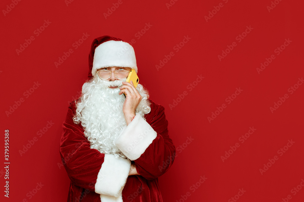 Portrait of a funny santa stands on a red background and talks on the phone with a serious face. Santa is calling on smartphone. Isolated. Christmas concept. Copyspace Portrait of a funny santa stands on a red background and talks on the phone with a serious face. Santa is calling on smartphone. Isolated. Christmas concept. Copyspace