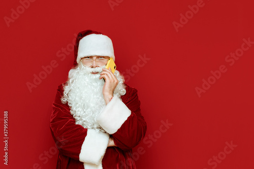 Portrait of a funny santa stands on a red background and talks on the phone with a serious face. Santa is calling on smartphone. Isolated. Christmas concept. Copyspace © bodnarphoto