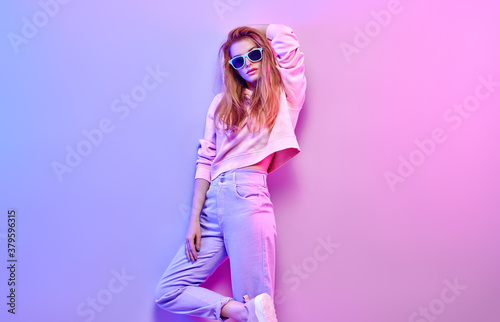 Fashionable hipster woman in Trendy outfit, stylish hair, makeup on pink purple neon light. Redhead model in hoodie, fashion jeans, sunglasses. Beautiful bad sexy girl in neon style