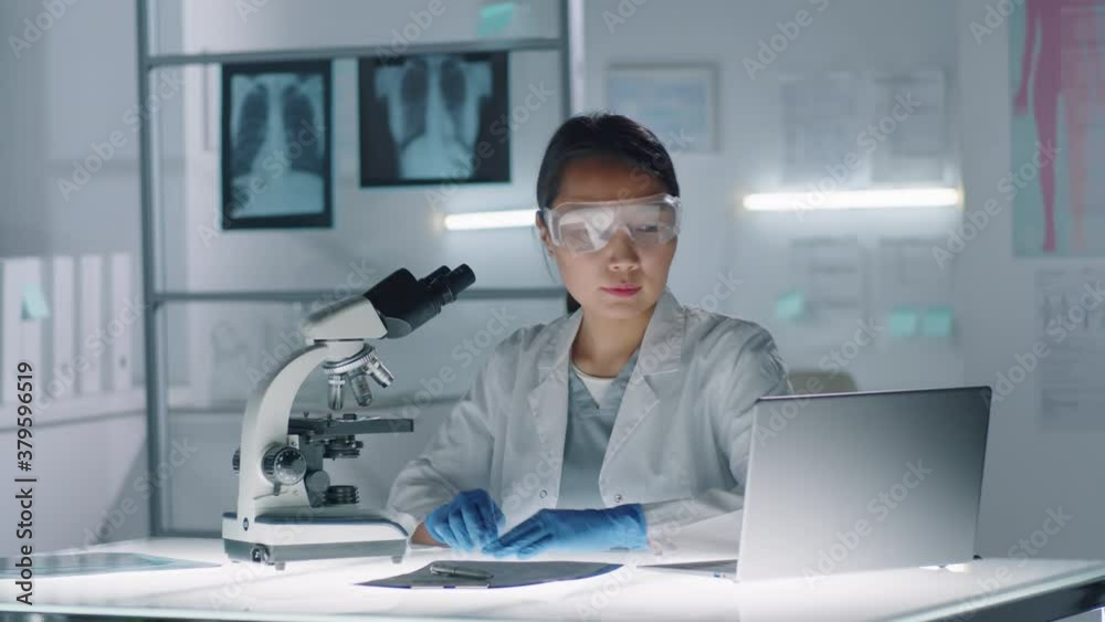 Slow motion footage of Asian woman wearing white coat, protective gloves and eyewear testing sample using microscope and laptop