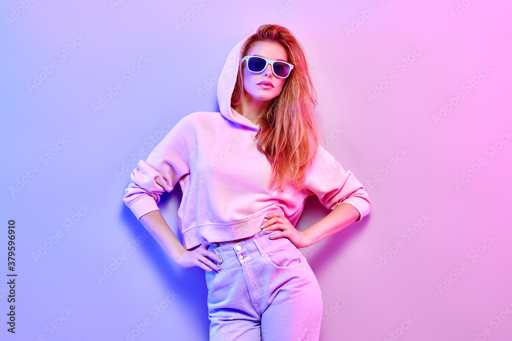 Fashionable hipster woman in Trendy outfit, stylish hair, makeup on pink purple neon light. Redhead model in hoodie, fashion jeans, sunglasses. Beautiful girl in autumn fall neon style