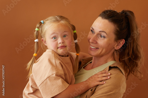 Waist up portrait of happy mature mother holding daughter with downs syndrome while standing against plain brown background in studio