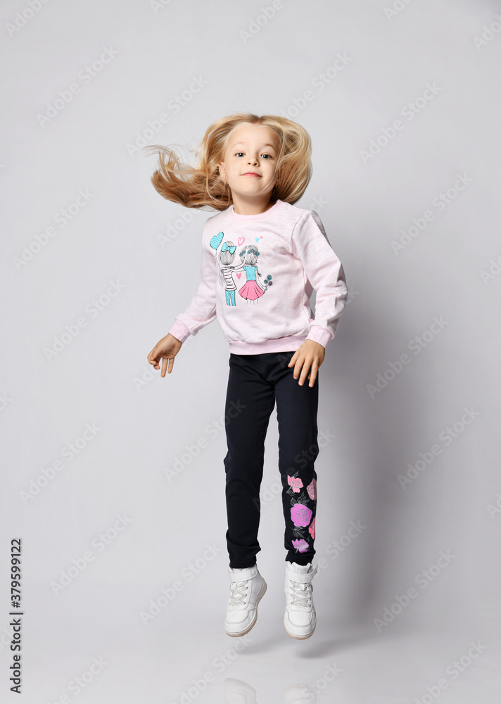 Cheerful blonde kid girl in gray sweatshirt sweater, blue pants and sneakers jumps Cheerful blonde kid girl in gray sweatshirt sweater, blue pants and sneakers jumps