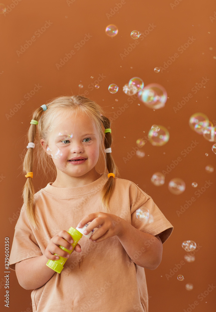 Verticalw aist up portrait of cute girl with down syndrome blowing bubbles while posing against brown background in studio