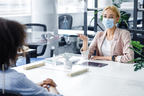 African american lady speaks to business woman in protective mask through glass partition in office interior