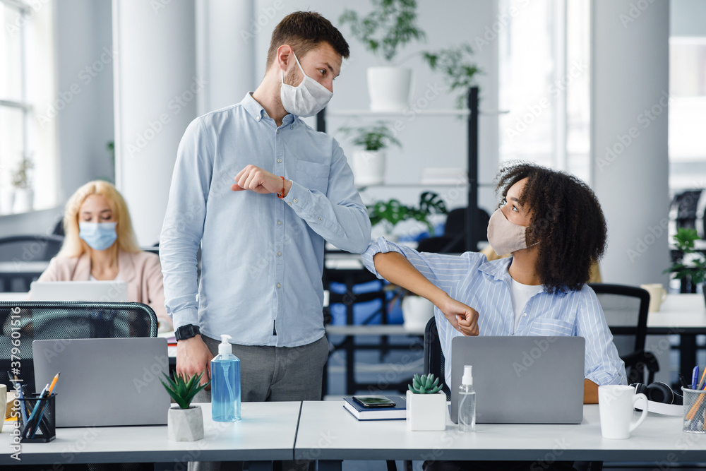 Fototapeta premium New normal and modern trend to say hello. Guy and african american lady in protective masks greet each other their elbows at workplace in interior of coworking office