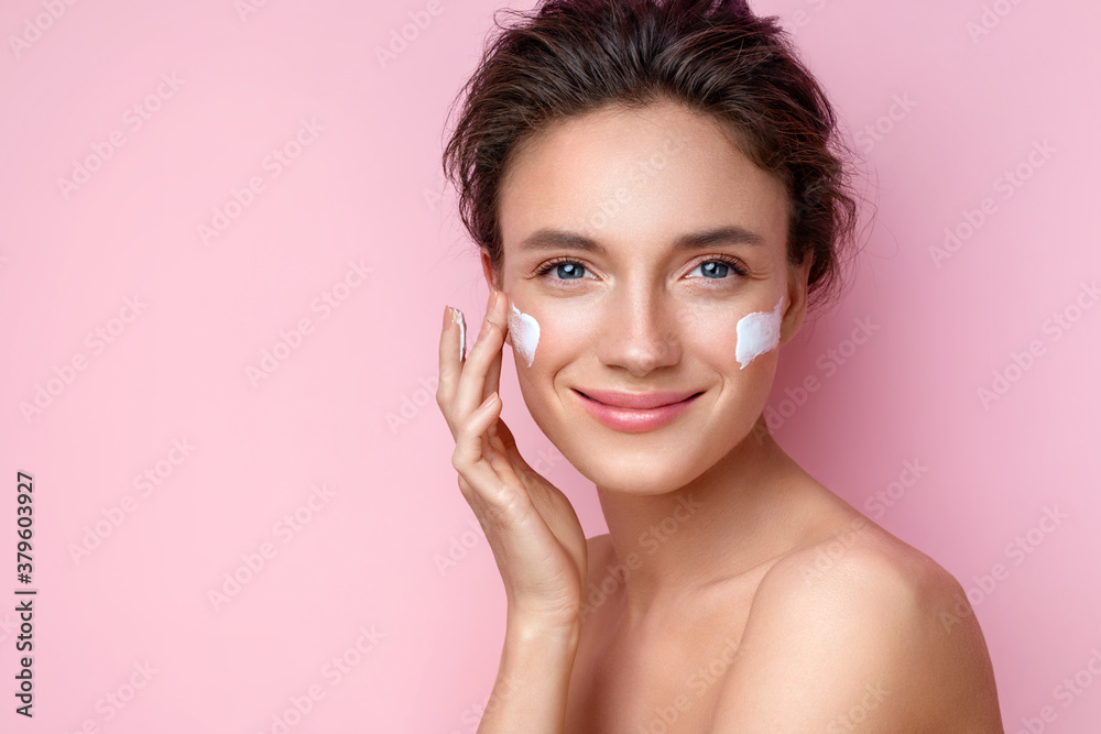 Beautiful woman applying moisturizer cream on her face. Photo of smiling woman with perfect makeup on pink background. Beauty concept - obrazy, fototapety, plakaty 
