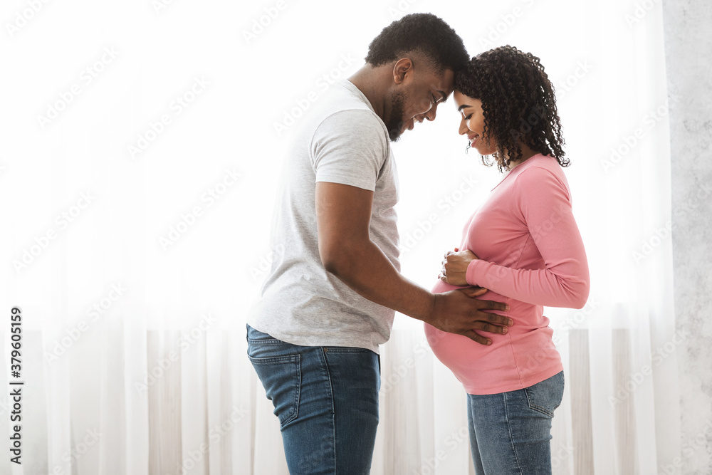 Black pregnant couple cuddling next to window at home