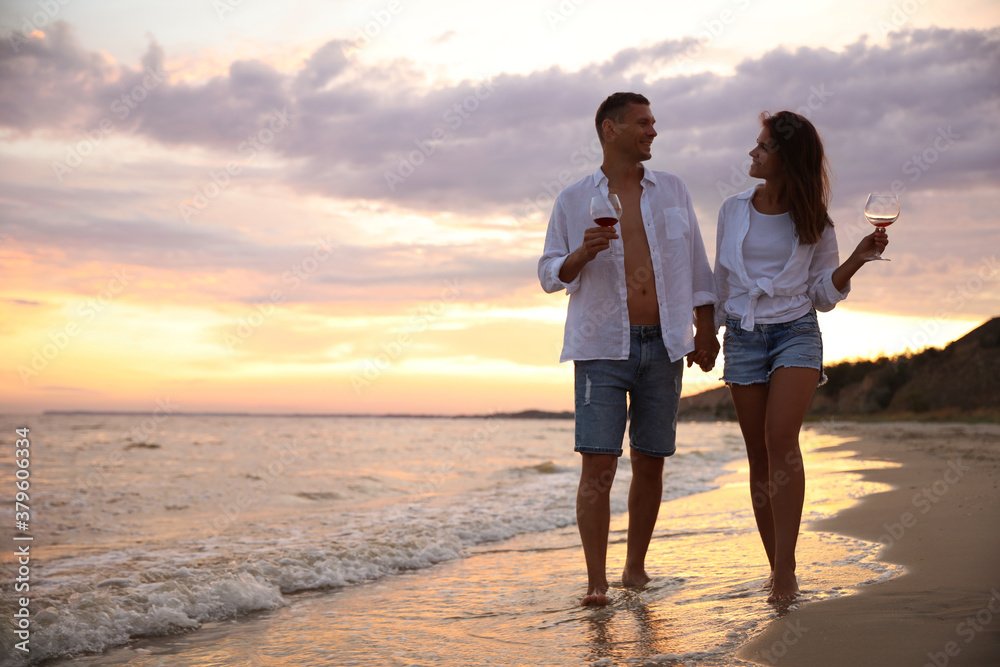 Lovely couple with glasses of wine walking on beach at sunset. Space for text