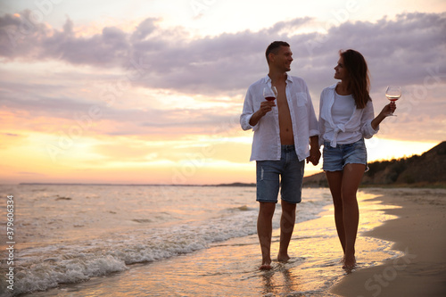 Lovely couple with glasses of wine walking on beach at sunset. Space for text