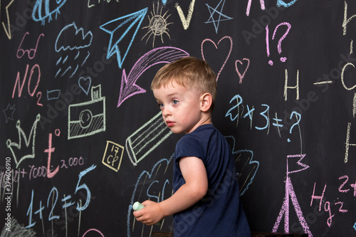 a little three-year-old boy stands near a large slate wall and draws drawings with colored chalk. European appearance.