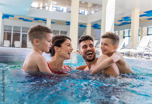 Smiling family of four having fun and relaxing in indoor swimming pool at hotel resort.