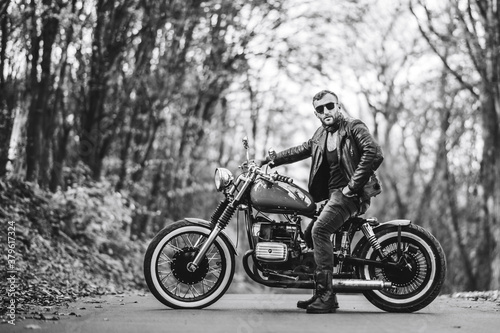 Bearded brutal man in sunglasses and leather jacket sitting on a motorcycle on the road in the forest © bedya