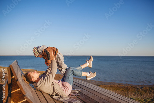 A young woman holds her baby at arm's length and laughs as she lies on a bench on the seaside in autumn