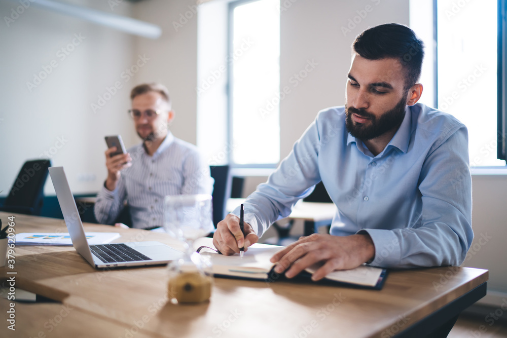 Caucasian man in formal t shirt writing strategy ideas in textbook sitting at table desktop with male colleague using smartphone gadget for phoning on blurred background, office working process