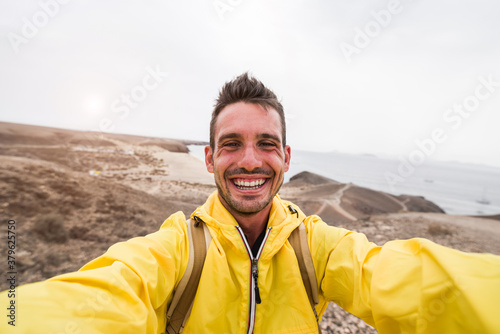 Handsome hiker man smiling at camera taking a selfie on the top of a mountain. People, sport, and technology concept.
