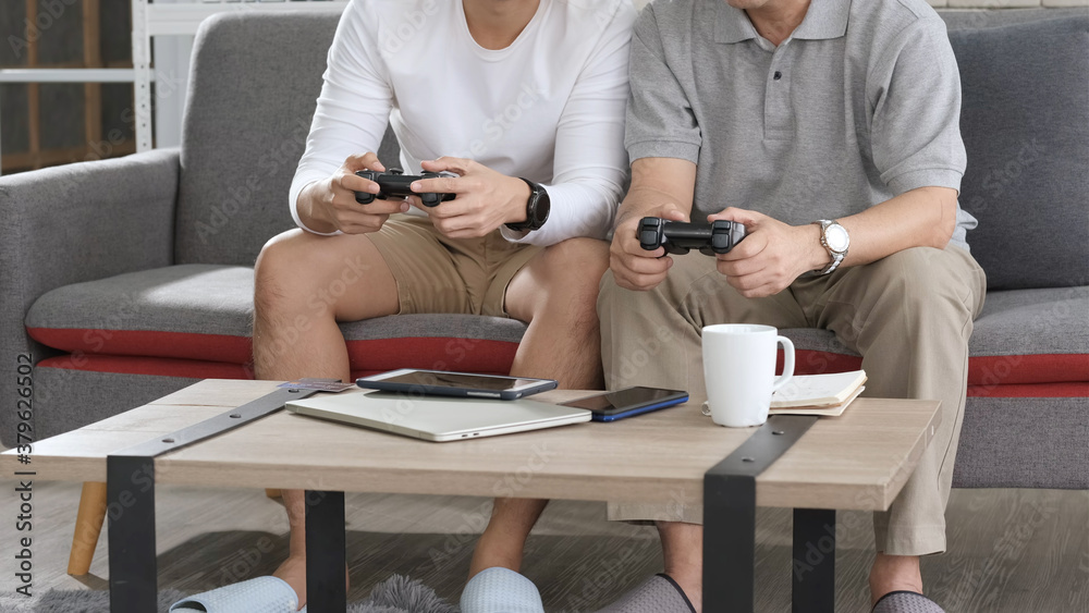 Fototapeta premium Asian two age generations men family old father embracing young adult son having fun enjoying play video game funny video using at home sit on sofa. Happiness Asian family concepts.