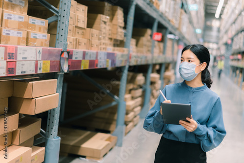 Young smart Asian business working woman wear surgical mask  using digital tablet to check goods on shelves for inventory management  in warehouse, Logistics business planning concept with copy space