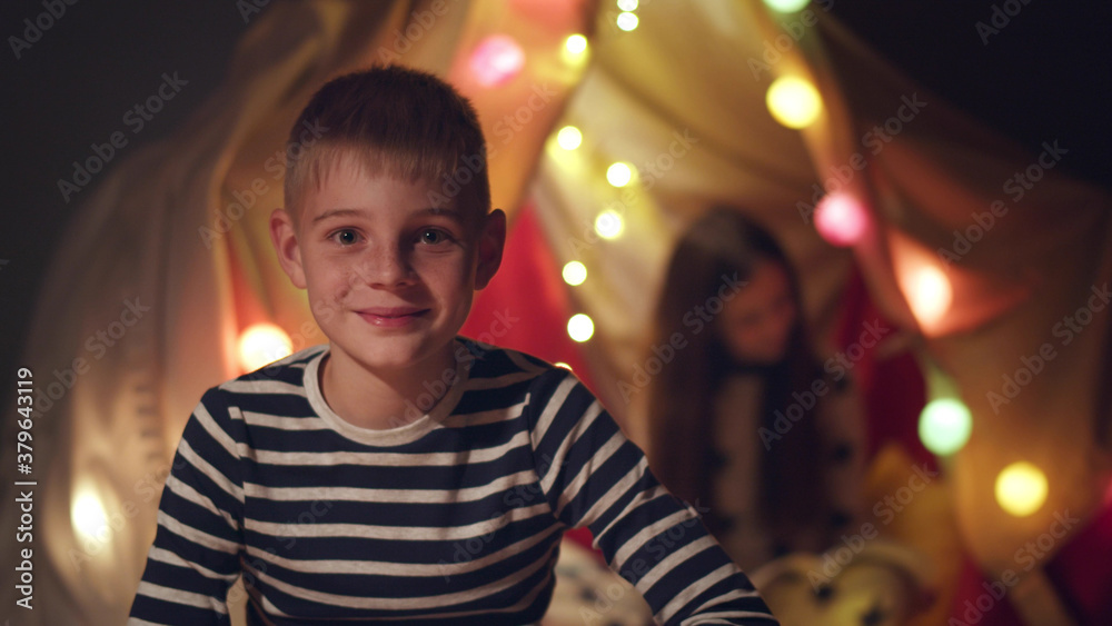 Happy smiling preteen boy in pajama smiling at camera with sister sitting in wigwam on background Happy smiling preteen boy in pajama smiling at camera with sister sitting in wigwam on background