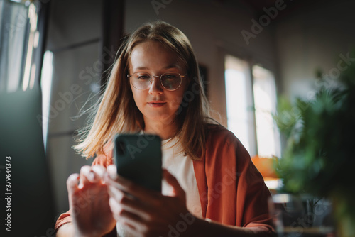 Young millennial girl in trnedy spectacles using smartphone while working remotely from home, Attractive woman typing on phone in modern loft, People Communication Technology photo