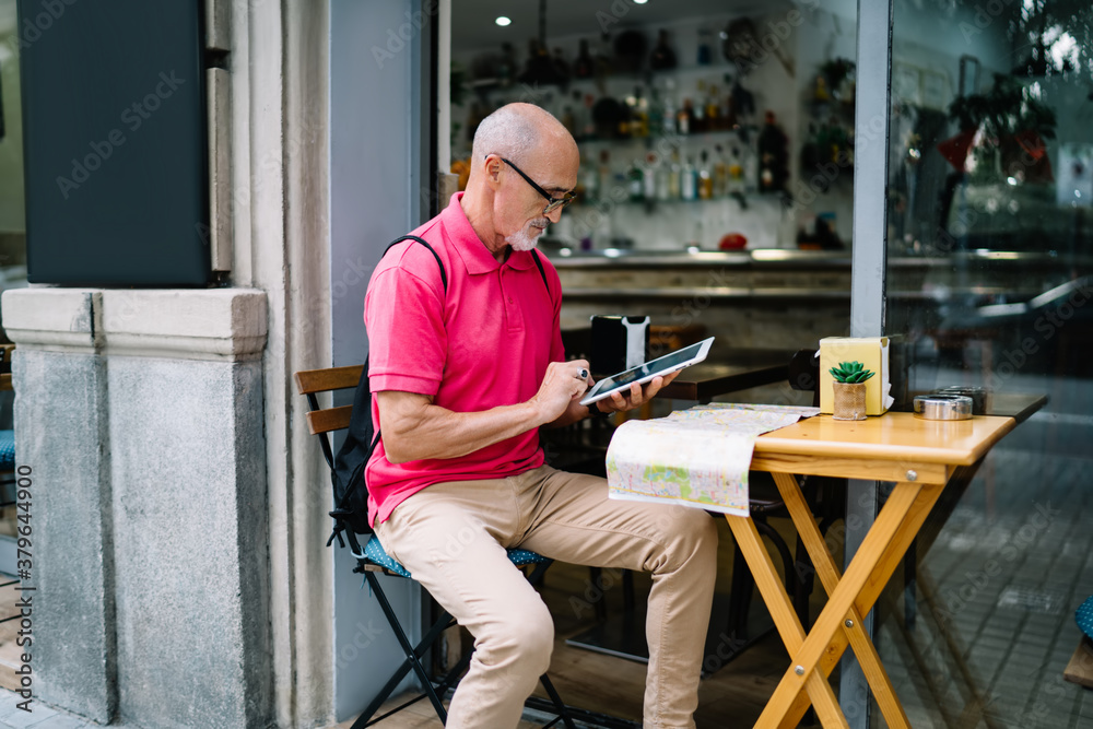 Mature male tourist connecting to 4g internet for using touch pad application and make online booking, man 60s in optical eyewear searching information via digital tablet sitting at sidewalk table