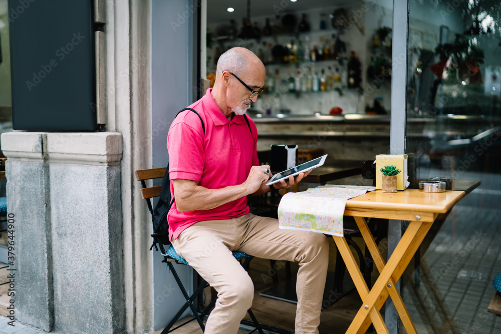 Mature male tourist connecting to 4g internet for using touch pad application and make online booking, man 60s in optical eyewear searching information via digital tablet sitting at sidewalk table Mature male tourist connecting to 4g internet for using touch pad application and make online booking, man 60s in optical eyewear searching information via digital tablet sitting at sidewalk table