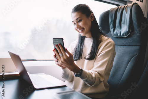 Cheerful female blogger working during trip on railroad sitting in train with wireless internet connection making picture on frontage mobile phone camera, happy woman posing for selfie on smartphone