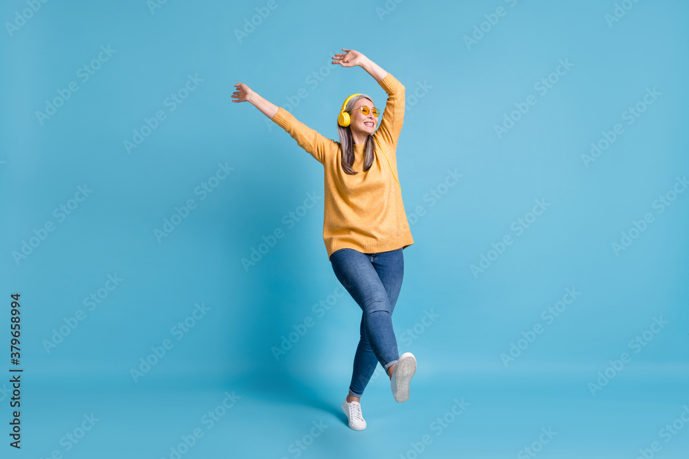 Full body size photo of attractive careless old dancer lady listen music earphones hands waving above head walking street wear sunglass yellow jumper jeans vivid blue color background Full body size photo of attractive careless old dancer lady listen music earphones hands waving above head walking street wear sunglass yellow jumper jeans vivid blue color background