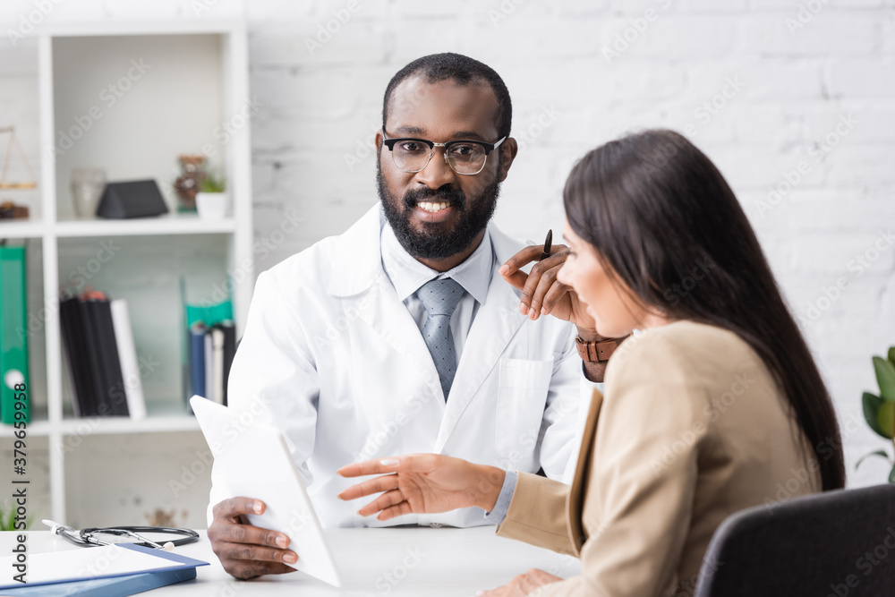 african american doctor holding digital tablet and looking at camera near brunette woman pointing with finger african american doctor holding digital tablet and looking at camera near brunette woman pointing with finger