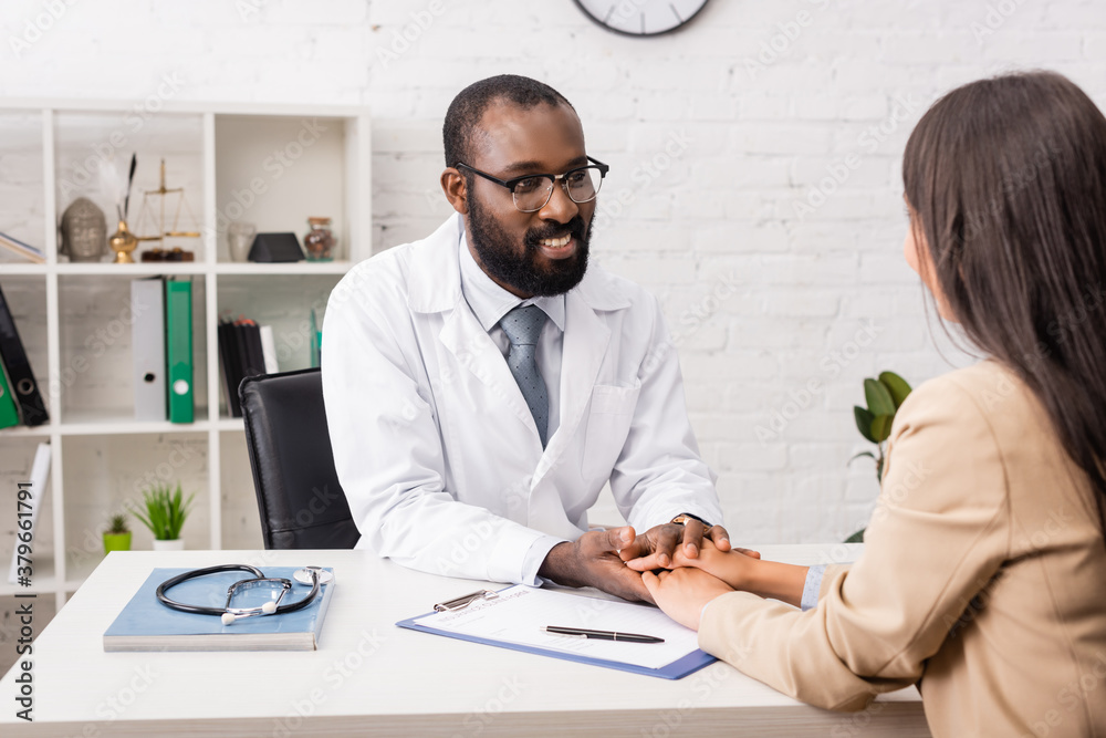 african american doctor in eyeglasses touching hands of brunette patient near clipboard with insurance form african american doctor in eyeglasses touching hands of brunette patient near clipboard with insurance form