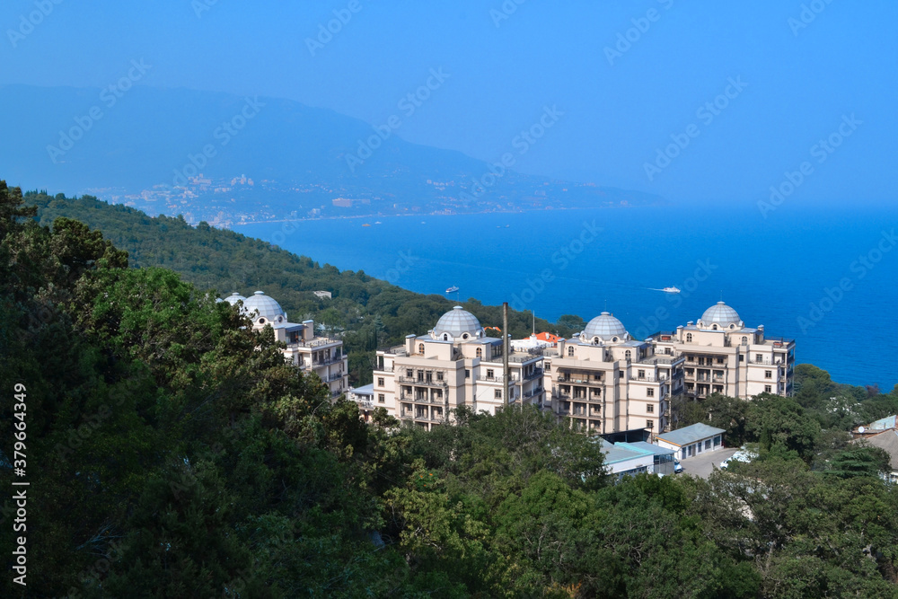 top view of resort town with white buildings on shore of Black Sea bay among mountains, blue water, summer, green forest, trees in the foreground