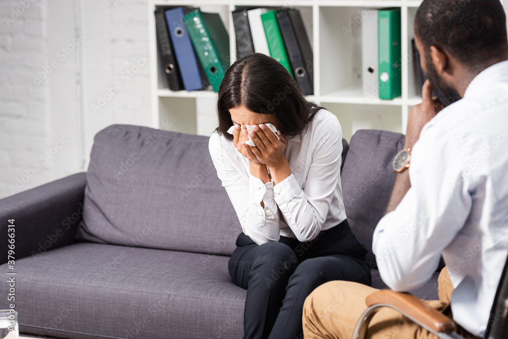 selective focus of african american psychologist sitting near crying woman wiping eyes with paper napkin selective focus of african american psychologist sitting near crying woman wiping eyes with paper napkin