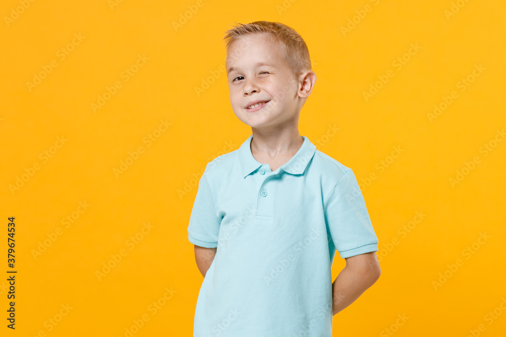 Smiling handsome little fun male fair-haired brown-eyed kid boy 5-6 years old wearing stylish blue turquoise t-shirt polo winking eye isolated on yellow color wall background, child studio portrait. Smiling handsome little fun male fair-haired brown-eyed kid boy 5-6 years old wearing stylish blue turquoise t-shirt polo winking eye isolated on yellow color wall background, child studio portrait.