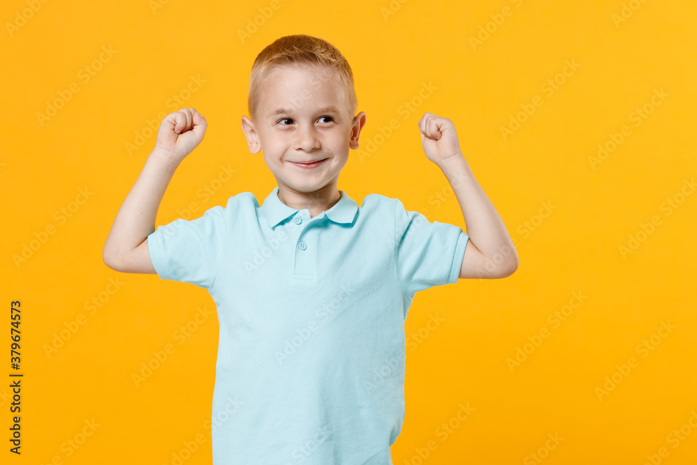 Smiling happy little fun male fair-haired brown-eyed kid boy 5-6 years old wearing stylish blue turquoise t-shirt polo raised hands up isolated on yellow color wall background, child studio portrait. Smiling happy little fun male fair-haired brown-eyed kid boy 5-6 years old wearing stylish blue turquoise t-shirt polo raised hands up isolated on yellow color wall background, child studio portrait.