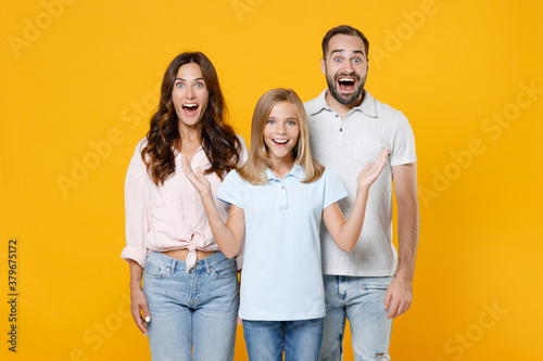 Surprised young parents mom dad with child kid daughter teen girl in basic t-shirts keeping mouth open spreading hands isolated on yellow background studio portrait. Family day parenthood concept.