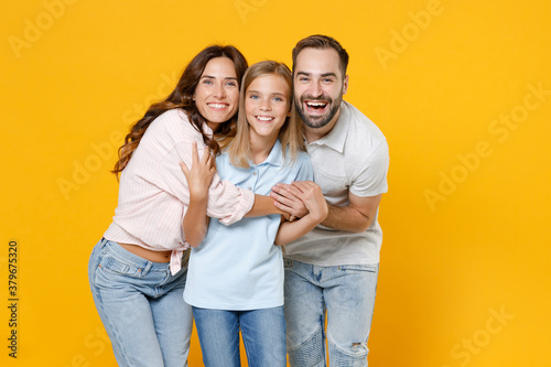 Laughing young happy parents mom dad with child kid daughter teen girl in basic t-shirts hugging looking camera isolated on yellow background studio portrait. Family day parenthood childhood concept.