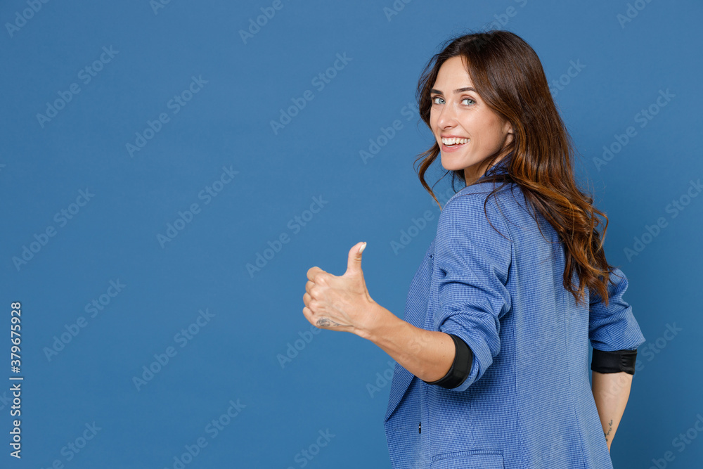 Back rear view of smiling cheerful fun attractive young brunette woman 20s wearing basic jacket standing looking camera showing thumb up isolated on bright blue colour wall background studio portrait. Back rear view of smiling cheerful fun attractive young brunette woman 20s wearing basic jacket standing looking camera showing thumb up isolated on bright blue colour wall background studio portrait.