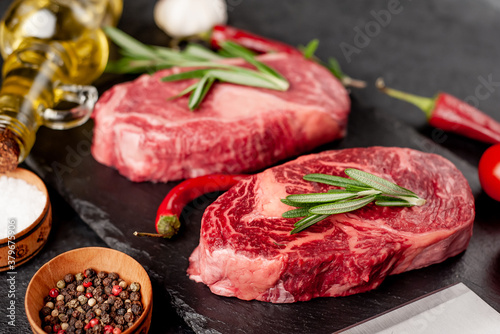 Two raw marbled beef ribeye steaks with spices on a stone background
