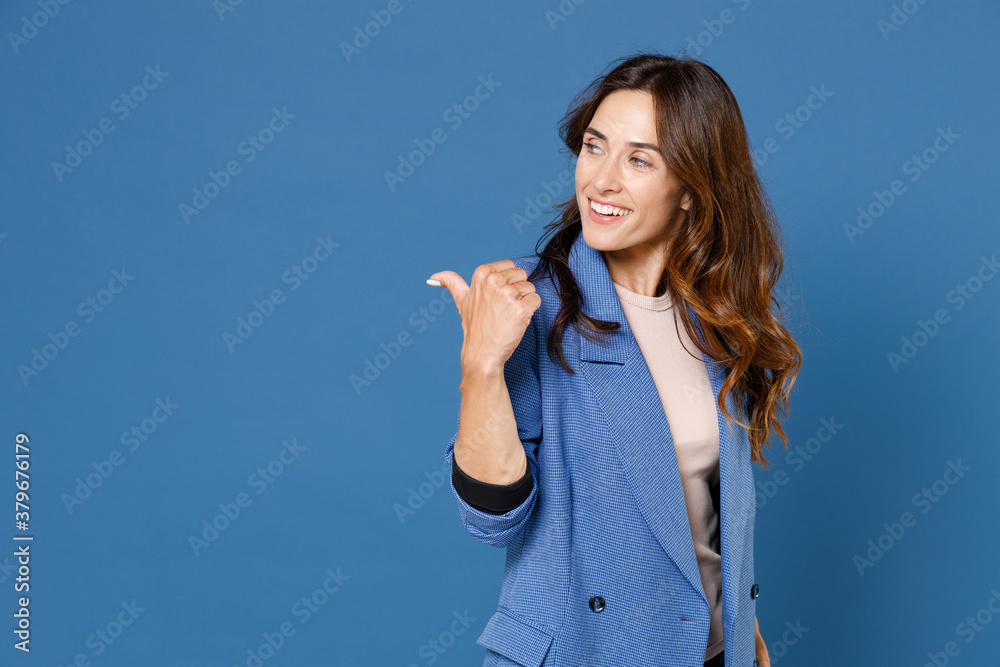 Smiling cheerful funny pretty beautiful young brunette woman 20s wearing basic jacket standing pointing thumb aside on mock up copy space isolated on bright blue colour background, studio portrait. Smiling cheerful funny pretty beautiful young brunette woman 20s wearing basic jacket standing pointing thumb aside on mock up copy space isolated on bright blue colour background, studio portrait.