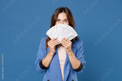 Fotografie Cheerful young brunette woman 20s wearing basic jacket standing covering face with fan of cash money in dollar banknotes looking camera isolated on bright blue colour wall background studio portrait